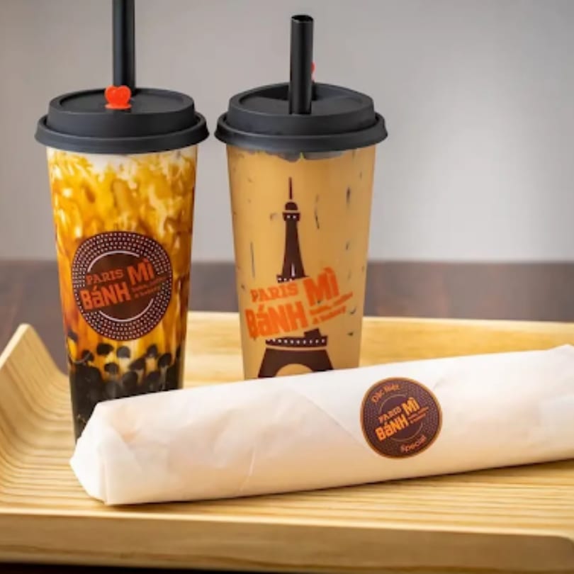 Two Paris Bánh Mì drinks and a wrapped bánh mì sandwich on a wooden tray.