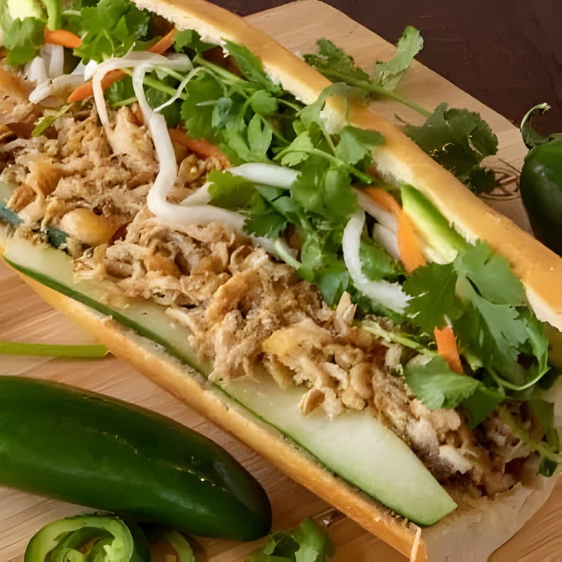 A Vietnamese bánh mì sandwich with crispy fried onions, fresh herbs, and sliced cucumbers on a wooden board.
