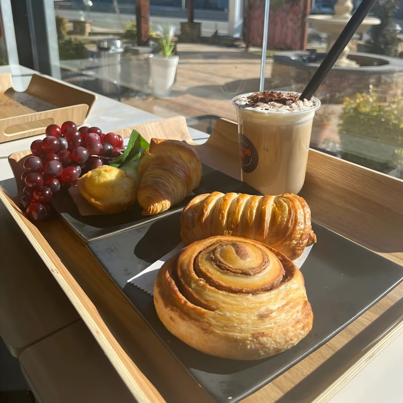 A wooden tray with pastries, red grapes, and an iced coffee with whipped cream by a window.