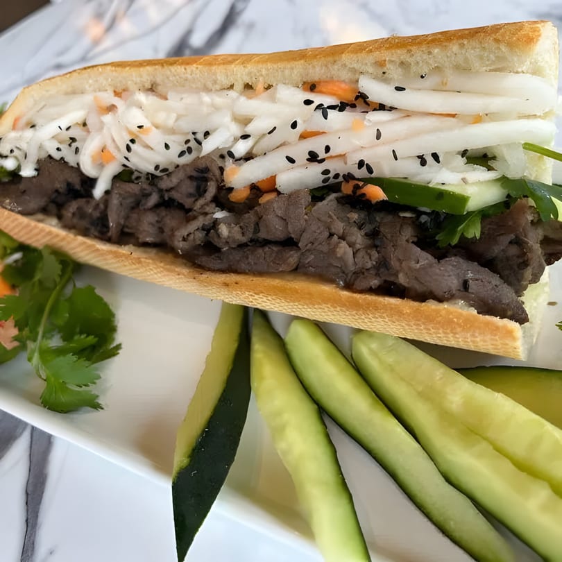 A Vietnamese Bánh Mì sandwich with beef, pickled daikon, carrots, and cucumbers on a white plate.